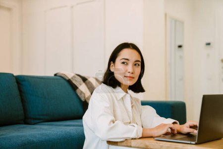 Téléchargez les photos : Jeune femme asiatique sérieuse mignonne avec ordinateur portable travaillant dans un salon confortable à la maison - en image libre de droit