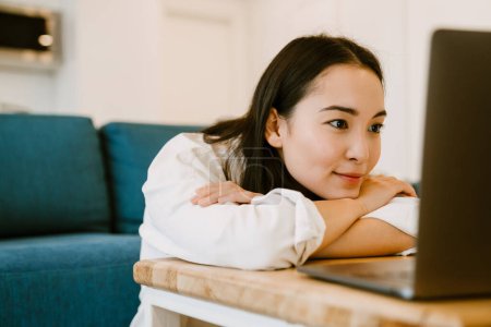 Téléchargez les photos : Jeune femme asiatique calme mignon avec ordinateur portable regardant sur l'écran dans le salon confortable à la maison - en image libre de droit