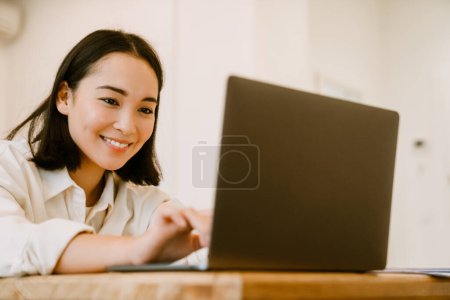 Téléchargez les photos : Jeune femme asiatique souriante mignonne avec ordinateur portable regardant à l'écran - en image libre de droit