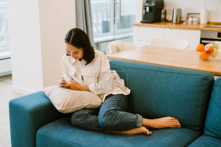 Foto de Joven linda chica asiática con teléfono de chat y sentado en el sofá en acogedora sala de estar espaciosa en casa - Imagen libre de derechos