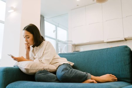 Téléchargez les photos : Jeune fille asiatique mignonne avec téléphone défilant les médias sociaux et assis sur le canapé dans le salon spacieux confortable à la maison - en image libre de droit