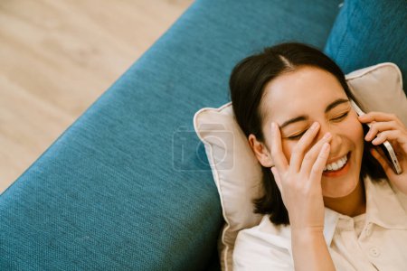 Téléchargez les photos : Jeune fille asiatique riante mignonne parlant au téléphone et couchée sur le canapé à la maison - en image libre de droit