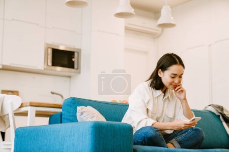 Photo for Young cute asian girl with phone looking on the screen sitting on couch in cozy spacious living room at home - Royalty Free Image