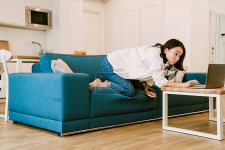 Foto de Joven hermosa sorprendida chica asiática llegar a la computadora portátil sentado en el sofá y en acogedora sala de estar espaciosa en casa - Imagen libre de derechos