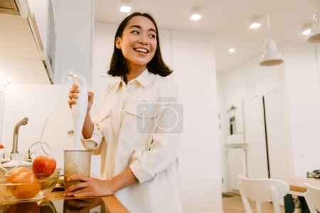 Téléchargez les photos : Jeune mignonne belle sourire asiatique fille cuisine smoothie dans la cuisine confortable à la maison - en image libre de droit