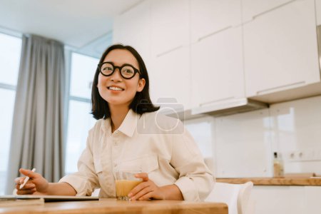 Téléchargez les photos : Jeune femme asiatique souriante mignonne dans des lunettes de travail par table dans la cuisine à la maison - en image libre de droit