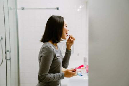 Téléchargez les photos : Jeune fille asiatique peignant ses lèvres avec un rouge à lèvres dans la salle de bain à la maison - en image libre de droit