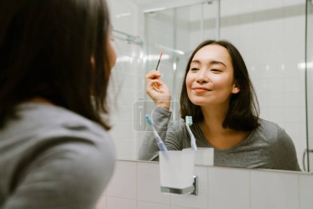 Téléchargez les photos : Jeune fille asiatique teindre les cils devant un miroir dans la salle de bain à la maison - en image libre de droit