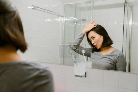 Téléchargez les photos : Jeune fille asiatique ajuster les cheveux devant un miroir dans la salle de bain à la maison - en image libre de droit