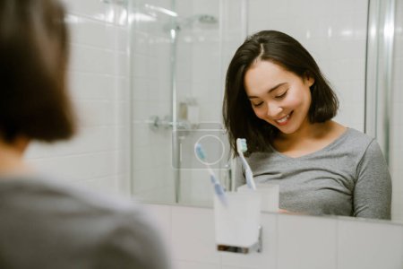 Téléchargez les photos : Jeune fille asiatique souriante debout devant un miroir et regardant vers le bas dans la salle de bain à la maison - en image libre de droit