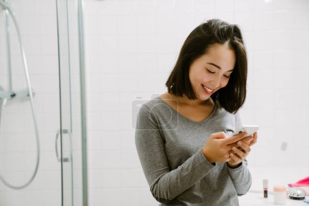 Photo for Young smiling asian girl with phone looking on screen in bathroom at home - Royalty Free Image