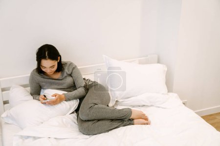 Photo for Young asian girl in home suit lying on the bed with phone at home - Royalty Free Image