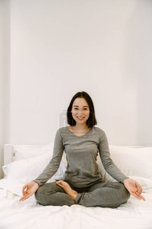 Photo for Young happy smiling asian girl in home suit sitting in lotus pose on the bed at home - Royalty Free Image