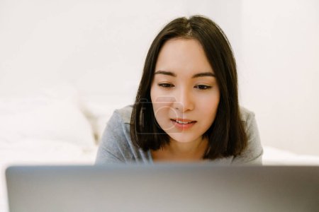 Téléchargez les photos : Jeune fille asiatique concentrée en costume à la maison reposant sur le lit avec ordinateur portable à la maison - en image libre de droit