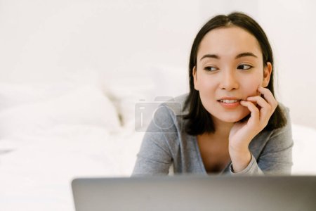 Téléchargez les photos : Jeune fille asiatique réfléchie en costume à la maison couché sur le lit avec ordinateur portable à la maison - en image libre de droit