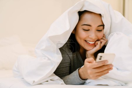 Téléchargez les photos : Jeune fille asiatique souriante en costume à la maison couché avec téléphone sur le lit sous les couvertures à la maison - en image libre de droit