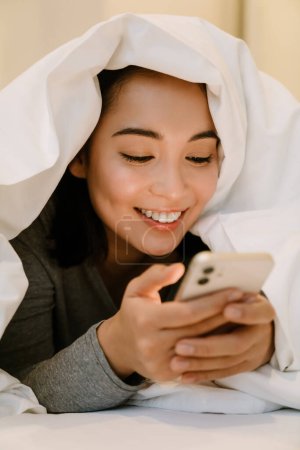 Foto de Joven sonriente chica asiática en traje de casa acostado con el teléfono en la cama debajo de las cubiertas en casa - Imagen libre de derechos