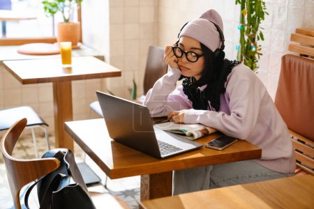 Téléchargez les photos : Fatigué asiatique femme dans casque faire des devoirs avec ordinateur portable au café - en image libre de droit