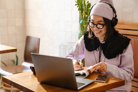 Téléchargez les photos : Jeune femme asiatique dans les écouteurs faire des devoirs avec ordinateur portable au café - en image libre de droit