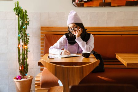 Foto de Joven asiático mujer en gafas de escribir notas mientras bebiendo café en la cafetería - Imagen libre de derechos