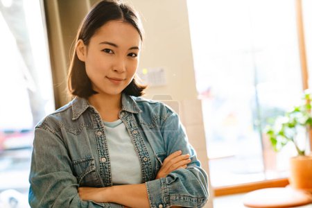 Téléchargez les photos : Jeune femme asiatique portant chemise en denim souriant et regardant la caméra à l'intérieur - en image libre de droit