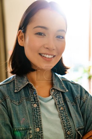 Téléchargez les photos : Jeune femme asiatique portant chemise en denim souriant et regardant la caméra à l'intérieur - en image libre de droit