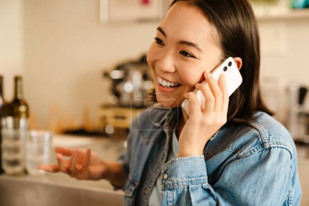 Téléchargez les photos : Jeune femme asiatique portant chemise en denim souriant et parlant sur téléphone portable au café - en image libre de droit