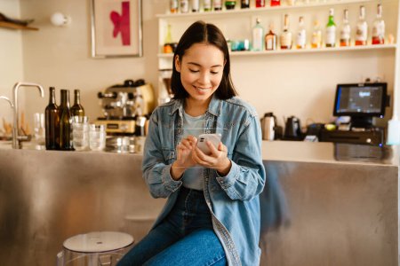 Téléchargez les photos : Jeune femme asiatique portant chemise en denim souriant et utilisant le téléphone mobile au café - en image libre de droit