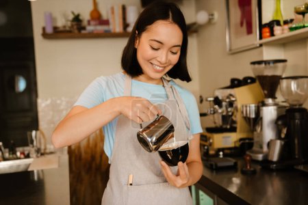 Téléchargez les photos : Jeune asiatique barista femme portant tablier souriant tout en faisant du café dans le café - en image libre de droit