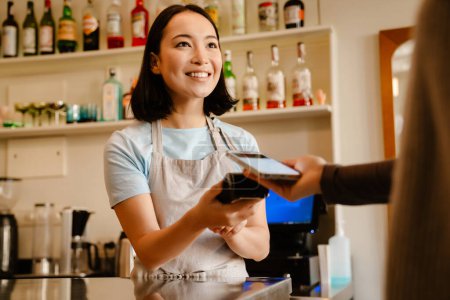 Asiatische Barista Frau trägt Schürze lächelnd, während sie mit Kunden im Café arbeitet
