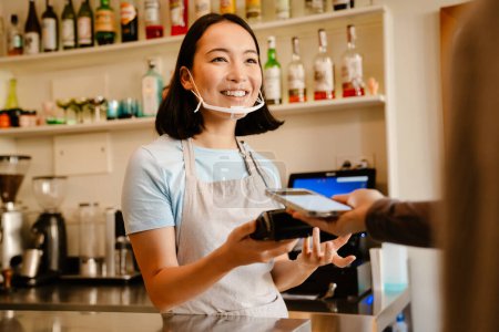 Photo for Asian barista woman in face mask smiling while working with client in cafe - Royalty Free Image