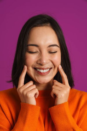 Photo for Asian young woman laughing while pointing fingers at her smile isolated over purple background - Royalty Free Image