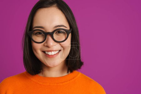 Foto de Joven mujer asiática en gafas sonriendo y mirando a la cámara aislada sobre fondo púrpura - Imagen libre de derechos