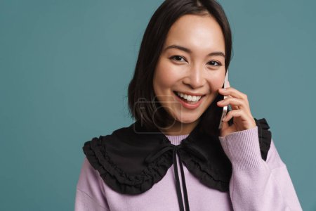 Téléchargez les photos : Jeune femme asiatique souriant et parlant sur téléphone mobile isolé sur fond bleu - en image libre de droit