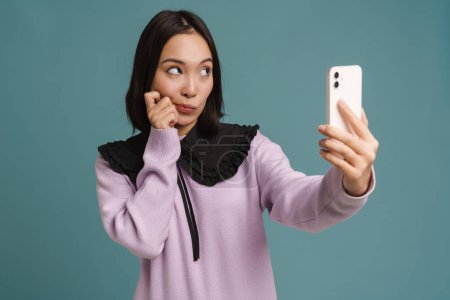 Photo for Young asian woman looking aside while taking selfie on cellphone isolated over blue background - Royalty Free Image