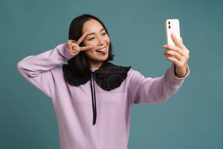 Photo for Young asian woman gesturing while taking selfie on cellphone isolated over blue background - Royalty Free Image
