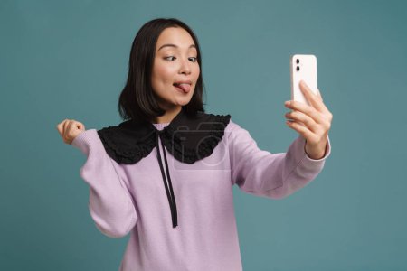 Photo for Young asian woman showing her tongue while taking selfie on cellphone isolated over blue background - Royalty Free Image