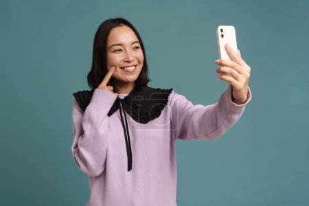 Photo for Young asian woman pointing finger at her cheek while taking selfie photo isolated over blue background - Royalty Free Image