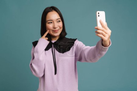 Photo for Young asian woman pointing finger at her cheek while taking selfie photo isolated over blue background - Royalty Free Image