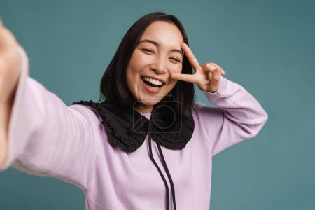 Téléchargez les photos : Jeune femme asiatique geste tout en prenant selfie photo isolé sur fond bleu - en image libre de droit