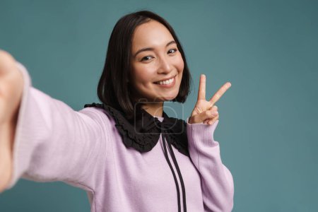 Photo for Young asian woman gesturing while taking selfie photo isolated over blue background - Royalty Free Image