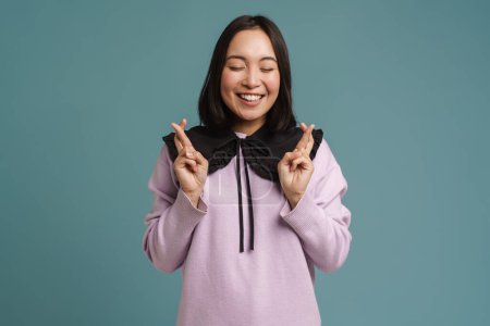 Photo for Young asian woman laughing while fingers crossed for good luck isolated over blue background - Royalty Free Image