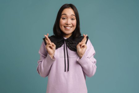 Téléchargez les photos : Jeune femme asiatique riant tandis que les doigts croisés pour la bonne chance isolé sur fond bleu - en image libre de droit