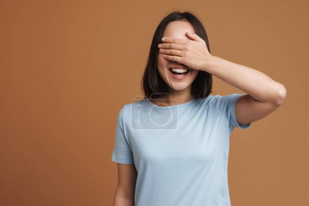 Téléchargez les photos : Jeune femme asiatique portant t-shirt riant et couvrant ses yeux isolés sur fond beige - en image libre de droit