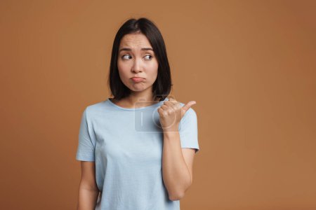 Photo for Perplexed asian woman wearing t-shirt pointing finger aside isolated over beige background - Royalty Free Image