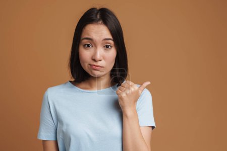 Photo for Perplexed asian woman wearing t-shirt pointing finger aside isolated over beige background - Royalty Free Image