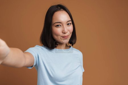 Photo for Young asian woman laughing while taking selfie photo isolated over beige background - Royalty Free Image