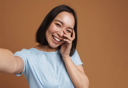 Foto de Joven mujer asiática riendo mientras toma foto selfie aislado sobre fondo beige - Imagen libre de derechos