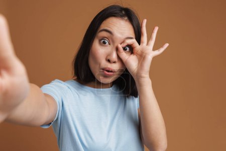 Téléchargez les photos : Surpris asiatique femme geste tout en prenant selfie photo isolé sur fond beige - en image libre de droit