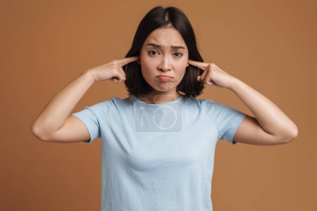 Téléchargez les photos : Jeune femme asiatique portant t-shirt fronçant les sourcils tout en bouchant ses oreilles isolées sur fond beige - en image libre de droit
